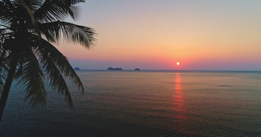 Beautiful sunset over the calm sea creating warm colors reflecting on the water surface with palm tree silhouette and islands on the horizon in Koh Samui, Thailand