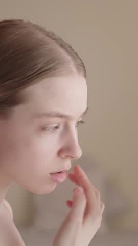 VERTICAL VIDEO: Young woman looks in the mirror, checking the condition of her facial skin. Gen Z female touching face with concern. Skin problems, aging, acne. Skincare products, dermatology services