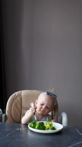 Independent toddler girl eating broccoli with spoon at table, sitting in high chair, practicing healthy eating and self feeding skills