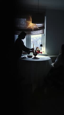 Light casts a warm glow as a person arranges a table with care, creating a welcoming atmosphere for guests in a serene indoor space. The room is beautifully adorned with festive decorations.