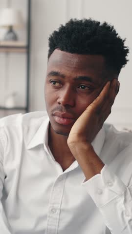 Portrait of bored african american man in room, vertical video, psychological problems. Emotional black male person thinking about troubles, startup owner or young businessman dreaming about money