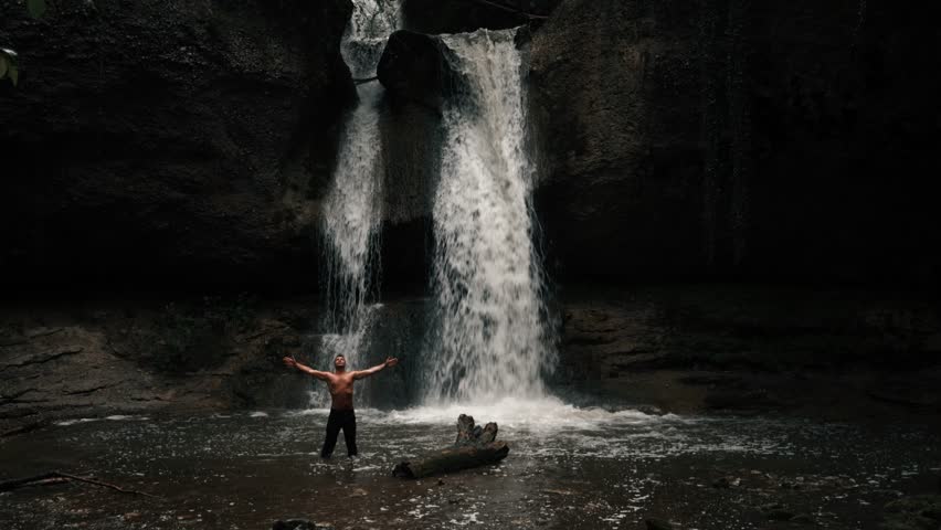 A man stands in the water in front of a waterfall in a mystical forest. It