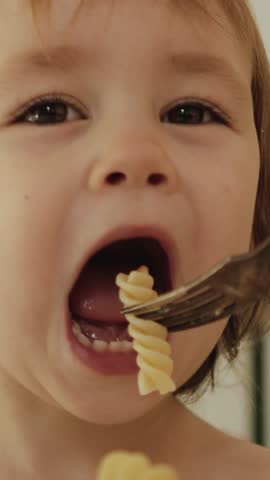 Vertical video. Portrait of happy child enjoying delicious food in bright kitchen, waving hands and smiling, eating pasta. Mother feeding her child at the table.