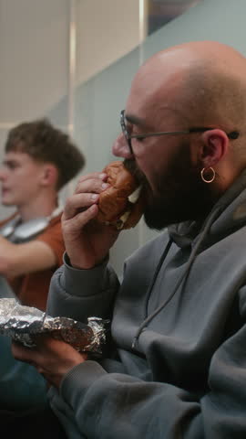 Diverse Passengers in Crowded International Airport Terminal: Man Eating Fast Food Burger while Sitting on the Floor in Departure Lounge with Suitcase and Waiting for Delayed Flight. Vertical Shot.