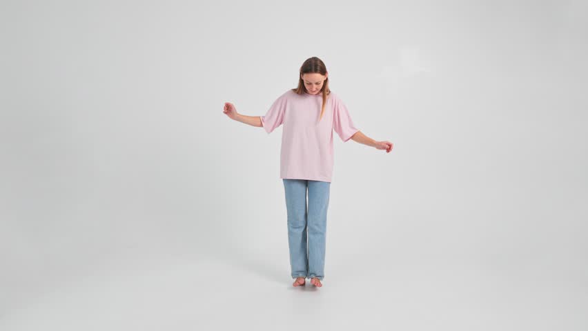 Woman in jeans and T-shirt is going on heels trying not to fall and holding hands in sides looking at the floor. Slow motion isolated footage of young adult Caucasian lady walking forward and back