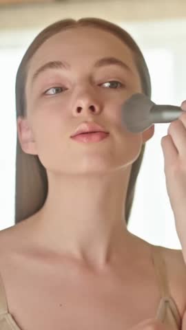 VERTICAL VIDEO: Beauty young woman applying makeup with a brush as part of her morning beauty routine. A happy Gen Z female enjoying self-care and preparing for the day. Confidence and beauty routine.
