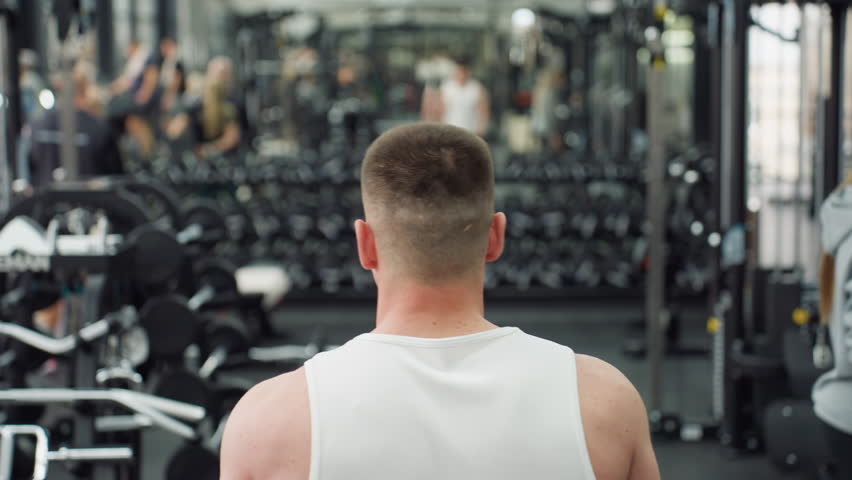 Shoulder back view of athlete strolling through athletic club filled with people exercising, rows of dumbbells and mirrors reflecting movement under lights, capturing energy and active atmosphere