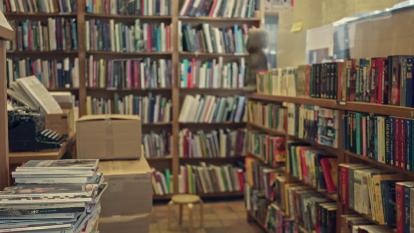Blurred library with bookshelves full of text books, magazines stacked, and cardboard boxes scattered indoors creating a cluttered academic atmosphere.