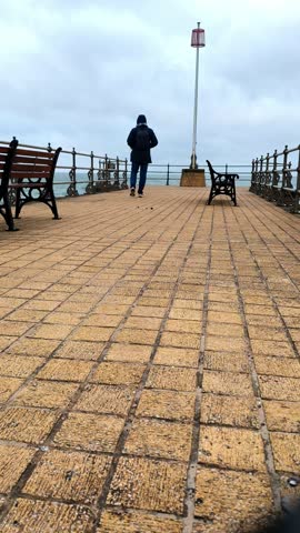 A man walks on a pier with a bench in the background. The bench is empty. The pier is made of bricks and the sky is cloudy