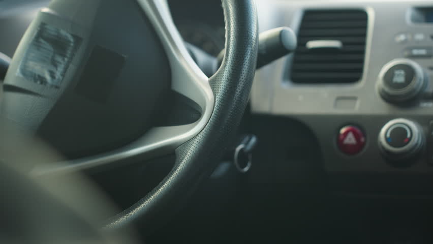 close up hand view of driver removing hand from ignition after turning on car, focus on steering wheel and dashboard area with climate controls and hazard button clearly visible