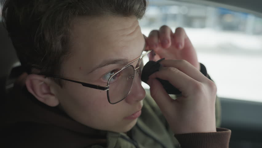 Young boy cleaning glasses lens with soft cloth inside car interior blur building visible through window under soft natural light and subtle reflections highlighting careful hand motion detail