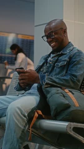 Crowded Airport Terminal: African American Man with Backpack Sitting in Departure Lounge After Passport Control, Scrolling Smartphone, Waiting for Flight. People at Security Check. Vertical Shot.