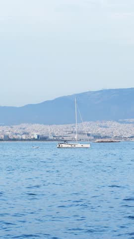 Serene twilight view of calm sea in Athens, Greece. A small white yacht in the distance, hazy mountains, faint cityscape, and soft spring colors in blue and white tones. No people. Extreme long shot
