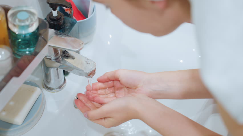 Lady gently washing her face at sink, water flowing from tap, skincare products visible beside sink, emphasizing facial cleansing and beauty routine for smooth, healthy skin