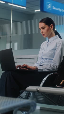 Airport Terminal: Successful Businesswoman Using Laptop, Doing e-Business or Remote Work, Looking at Camera. Entrepreneur Going on Business Trip, Waiting for Flight in Departure Lounge. Vertical Shot.