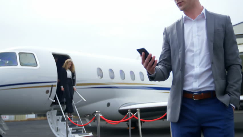 Businessman checking smartphone near jet by rope barrier on tarmac, with animated status chart. Luxury, professionalism, travel, corporate, elite, modern, sleek