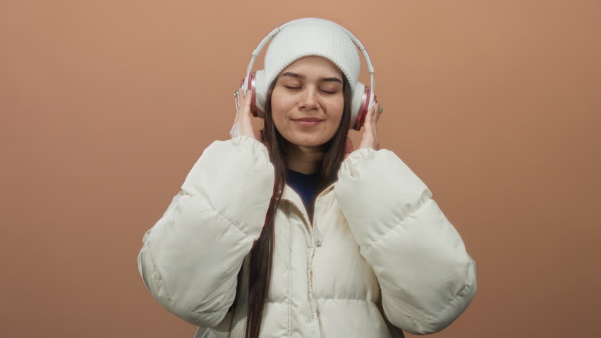Woman enjoying music with headphones wearing a white coat and beanie against an isolated brown background highlighting relaxation and style.