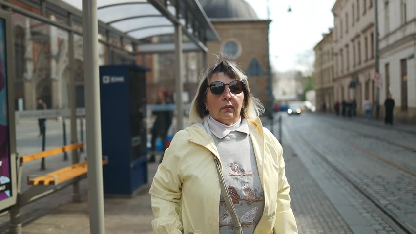 Elderly woman standing at a tram stop and looking around while waiting for public transport in Krakow, Poland. Urban travel scene highlighting mobility, independence, and city life for seniors.