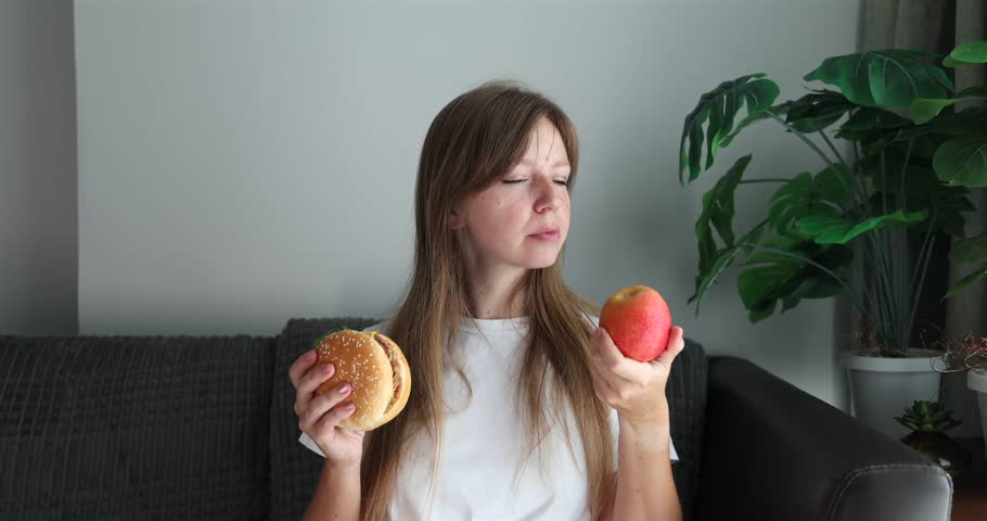 Woman deciding between burger and apple, healthy vs indulgence concept, nutrition choice, lifestyle and wellness theme.