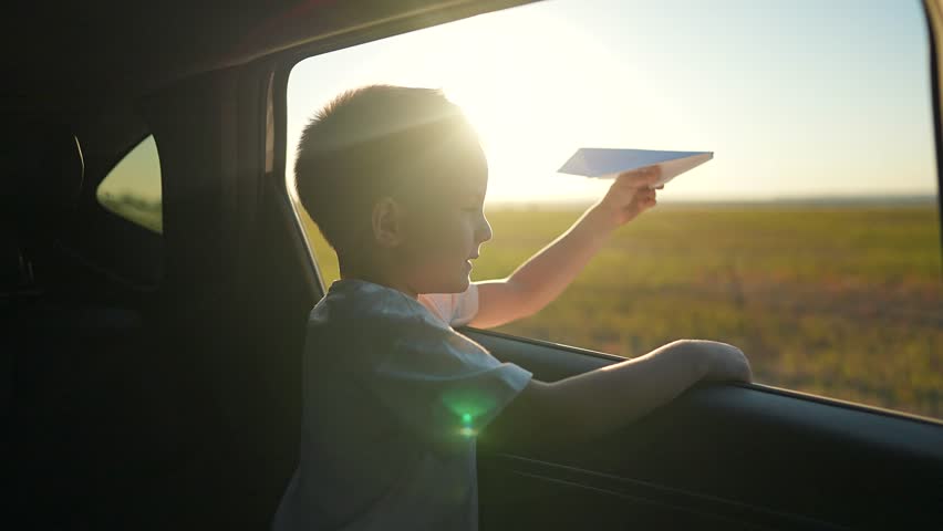 Child holding paper plane out car window. Kid enjoys playful moment with plane while traveling. Paper plane flying outside car window as child imagines adventure. child playing plane during road trip.