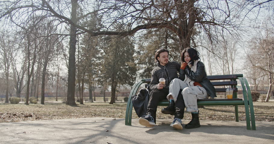 A happy couple enjoys a sunny winter day in the park, sitting on a bench, drinking coffee, and sharing a tender moment together.