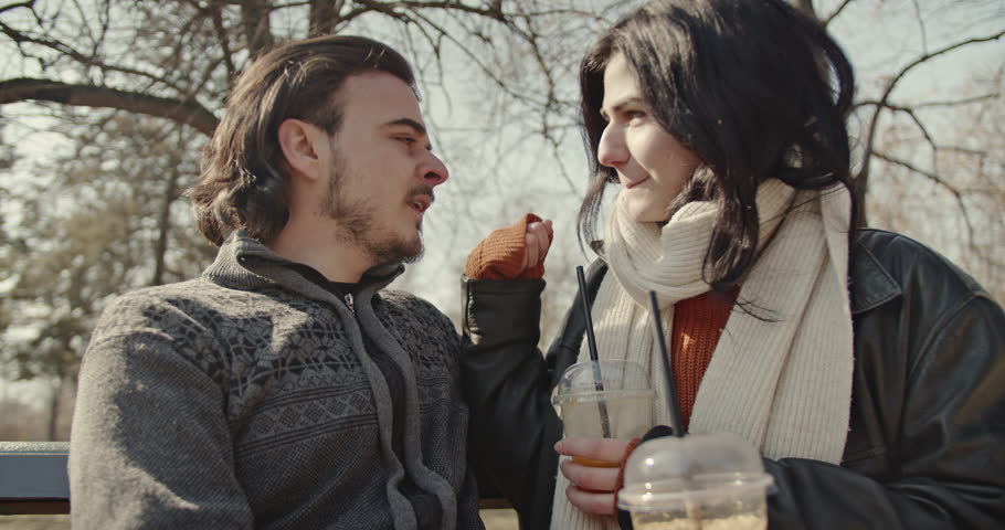 A happy couple enjoys a sunny winter day in the park. They sit on a bench, embrace, kiss, and sip drinks.