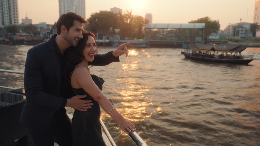 Portrait of loving Latin man shows interesting landmark to girlfriend sailing yacht at sunset. Hispanic couple enjoys romantic sea journey on vessel in Thailand. Date atmosphere