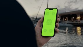 GREEN SCREEN CHROMA KEY person holding a smartphone with green screen near a marina with yachts and high-rise buildings in the background during sunset - Powered by Shutterstock - Get 15% off with code: PIKWIZARD15