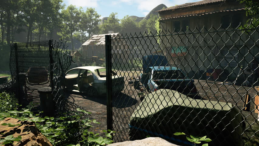 Through a chain-link fence, two stripped, weathered cars sit in a cluttered yard beside abandoned wooden building, surrounded by barrels, tools, and dense forest. Post-apocalyptic gaming setting.