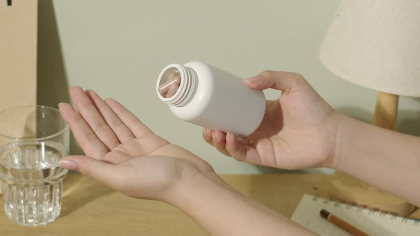 Hand holds the open drug bottle, tilts it sideways, shakes it gently and pours the medicine out so that the pills fall out. The brown capsule-shaped pills fall out of the bottle and onto the hand.