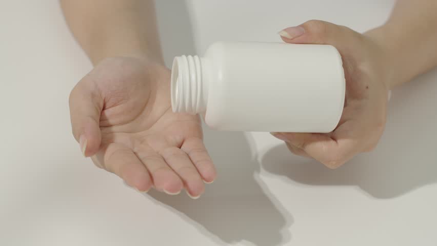 Close-up of hand holding pill bottle opening and pouring white pills into hand below. The compressed pills have a border in the middle to make it easy to break in half. Presented on white background.