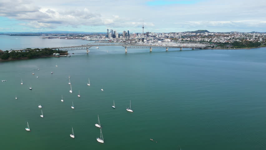 Drone flying toward Auckland Harbour Bridge with boats in Waitemata Harbour and city skyline in the distance, New Zealand.