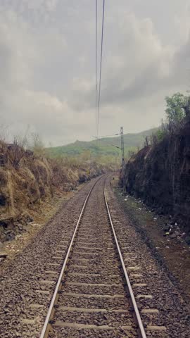 Train tracks stretch forward, flanked by gentle, grassy hills on both sides. The sky above is bright yet scattered clouds Maharashtra India 4K