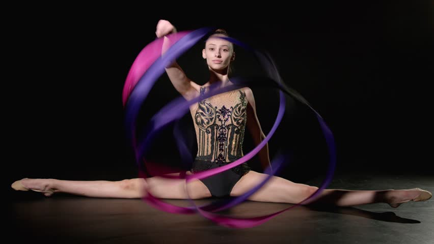 Flexible professional attractive rhythmic gymnast in elegant leotard sitting in side splits, performing spirals with bright ribbon against black background.