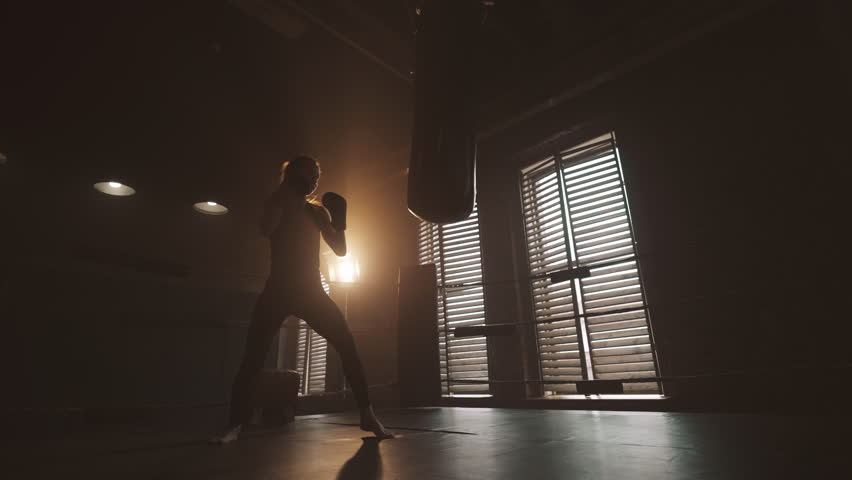 Intense cardio workout in boxing club, silhouette of slender lady and punch bag. Beautiful woman practicing punches by arms, hard training in sports gym, keeping fit and stay slender, slow motion