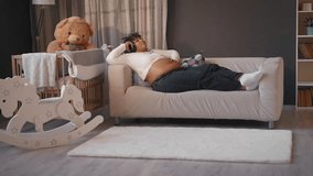 Pregnant woman relaxing in room and listening to classical music by headphones. African american lady enjoying relaxation and calmness, future mother lying on soft couch in bedroom, happy pregnancy - Powered by Shutterstock - Get 15% off with code: PIKWIZARD15