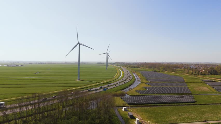 Wide aerial shot of Dutch countryside featuring renewable energy sources: tall wind turbines next to fields of solar panels along a modern highway. Captured in clear daylight.