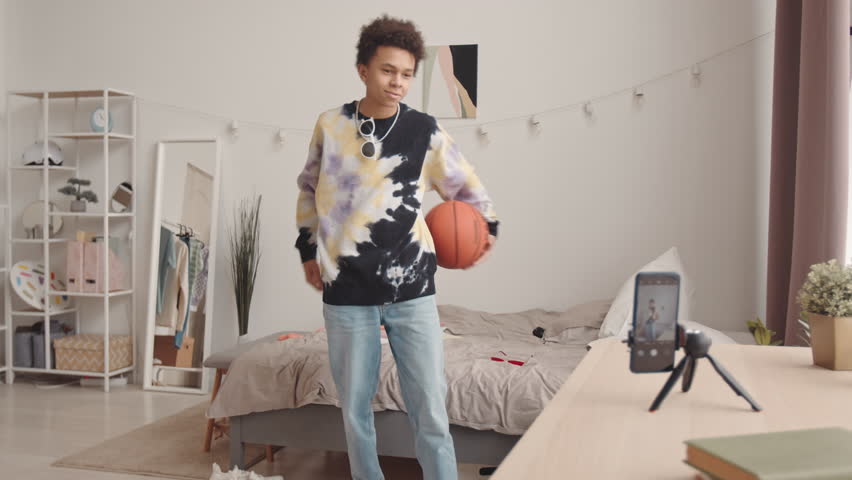 Slowmo of cool Biracial teenage boy making tricks with basketball and dancing while recording video blog on smartphone in his bedroom