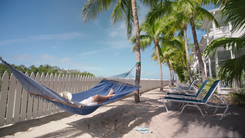 A woman is savoring a leisurely swing in slow motion on a hammock at a stunning hotel located in Key West, Florida, with palm trees surrounding her and breathtaking views all around her