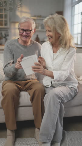 Aged people viewing old photographs, memories about happy life moments, vertical portrait in apartment. Happy aged spouses enjoying warm memories, smiling elderly people, calm and happy retirement