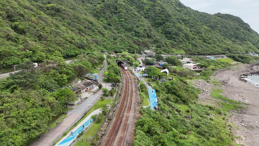 Aerial view of a Chu-Kuang Express train coming out of the tunnel to dash on the track between lush green mountains and the rocky beach by the Pacific Ocean on a sunny day, in Shicheng, Yilan, Taiwan