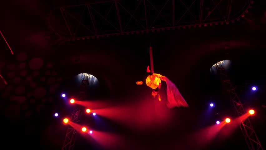 A flexible aerial gymnast performs tricks on fabric high under the dome of the circus under the light of colorful spotlights and smoke. A circus show. An aerial acrobat.