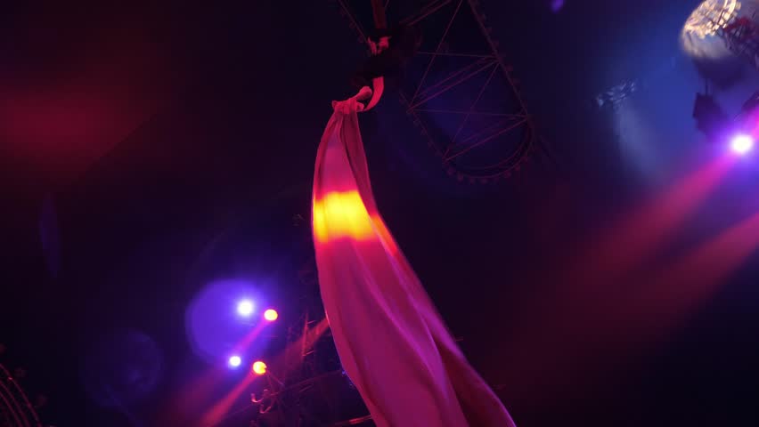 A flexible aerial gymnast performs tricks on the fabric high under the dome of the circus, she holds onto the fabric with her hands. Circus show. An aerial acrobat.