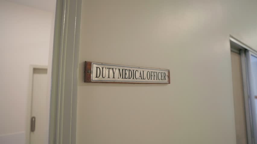 Sign reading "DUTY MEDICAL OFFICER" mounted beside a doorway on a cream-colored wall inside a hospital corridor, captured in a zoom-in movement with ambient daylight