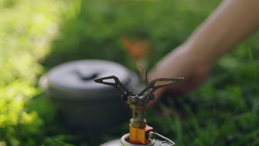 Person ignites portable gas stove with lighter. Cooking pot on burner. Outdoor cooking in nature. For camping, hiking, travel, eco-friendly trips, lightweight gear, off-grid adventures. Slow motion.