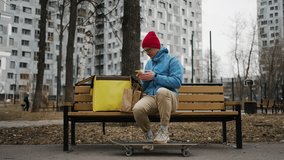 Food delivery man wearing thermal backpack sitting on bench with skateboard, checking order on smartphone, packs bags with orders and closing insulated bag before delivering food - Powered by Shutterstock - Get 15% off with code: PIKWIZARD15