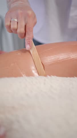 Beautician applying warm wax on client leg using wooden spatula during hair removal procedure