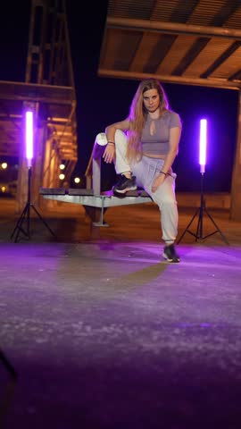 Confident young woman performing various hip hop dance moves on a rooftop at night, illuminated by blue neon lights