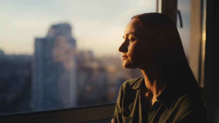 Lonely red-haired woman looking through window, feeling emotional and depressed