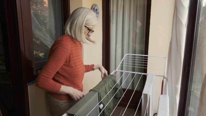 Senior woman wearing glasses arranging laundry on a drying rack indoors, near a sunny window, emphasizing daily chores and domestic lifestyles